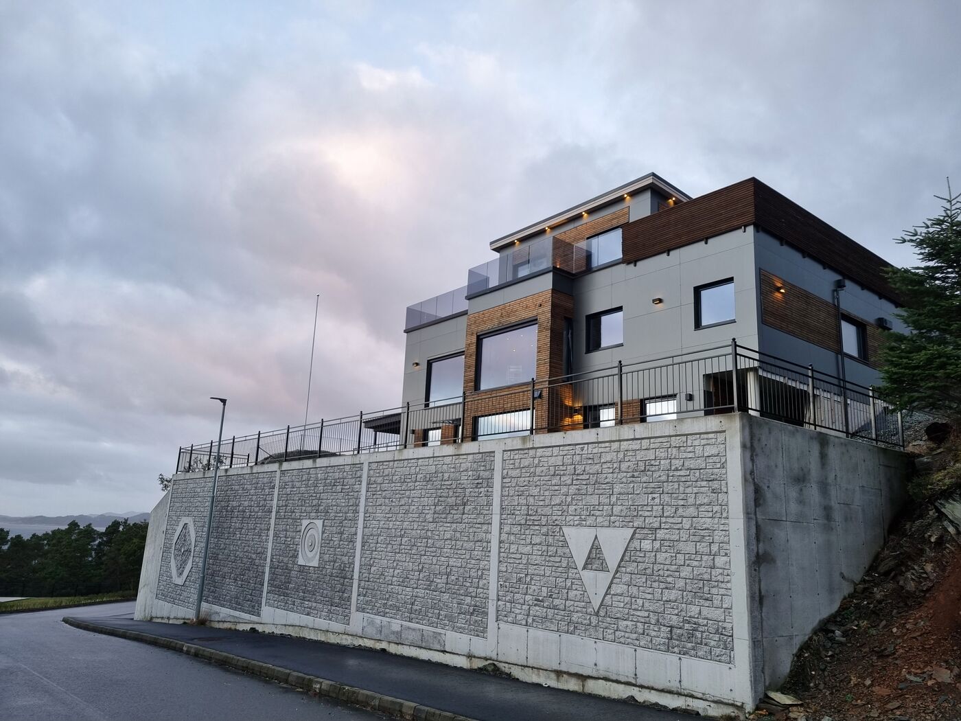 Huset sett fra veien med den plasstøpte støttemuren med relieffmønster, i kveldslys