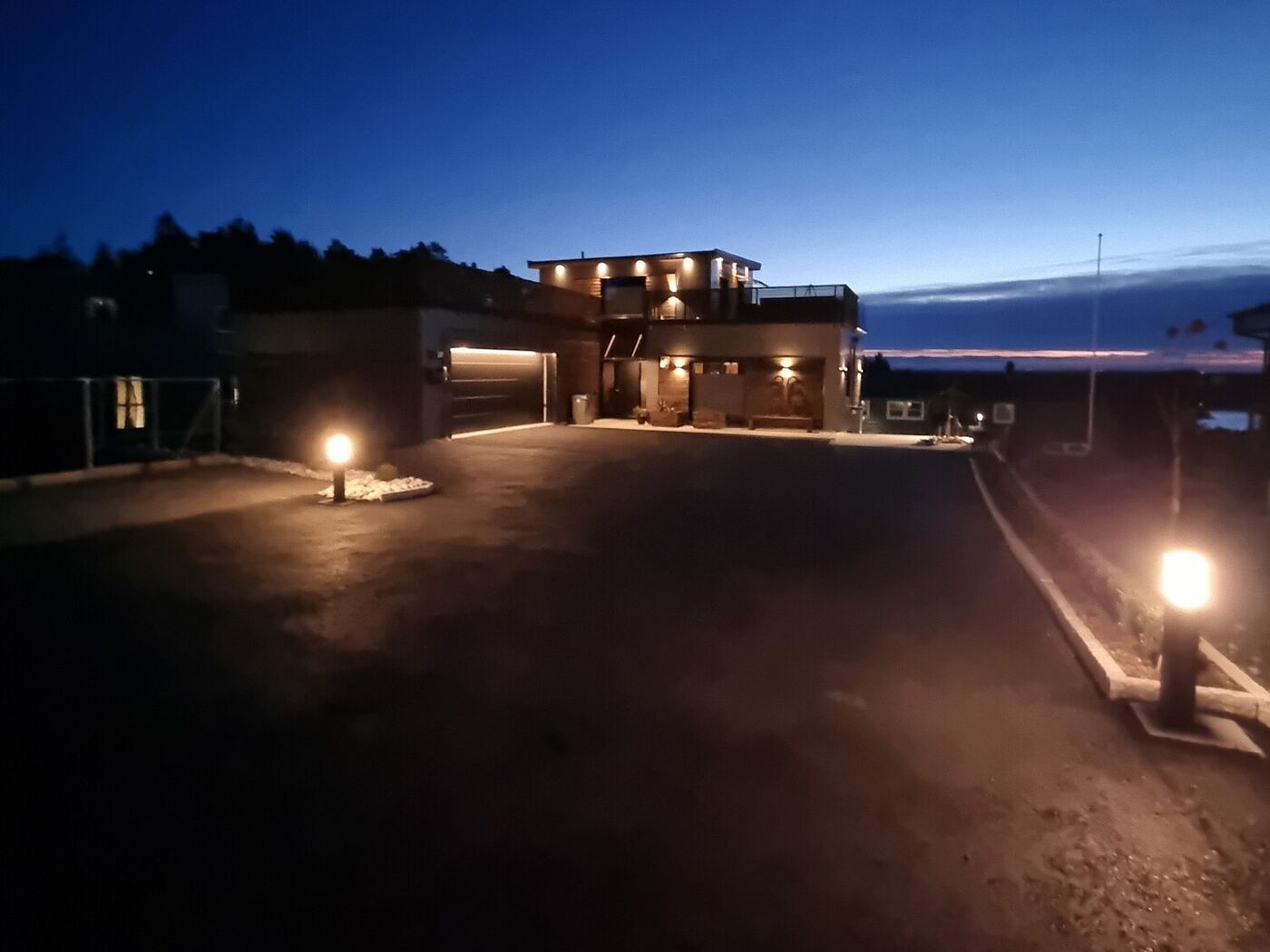 Eiendommen i blåtimen med opplyst innkjørsel, fasadebelysning og solnedgang i horisonten